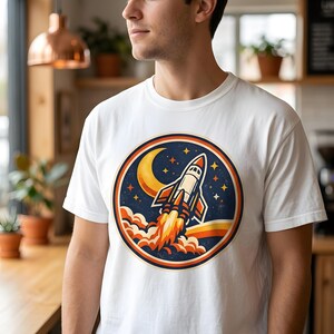 May include: White t-shirt featuring a circular graphic of a rocket ship blasting off into space. The design includes a crescent moon, stars, and a retro color palette of orange, yellow, and blue. The shirt is made of a soft, comfortable material.