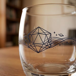 Puede incluir: Un vaso transparente sin pie con un diseño geométrico y acentos celestiales. El diseño azul oscuro presenta una forma multifacética, líneas onduladas, estrellas y un planeta. El vaso es probablemente para bebidas.