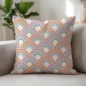 Op de afbeelding: Vierkant kussen met een herhalend patroon van regenboogbogen in bruin, paars, blauw en groen op een crèmekleurige achtergrond. Het kussen ligt op een grijze bank, geschikt voor interieurdecoratie.
