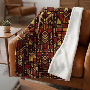 May include: A throw blanket with a geometric stained-glass design in shades of red, yellow, and black. The blanket is draped over a brown leather couch, with a soft white sherpa lining visible on the edge.