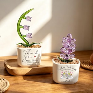 Può includere: Due vasi quadrati in ceramica bianca contenenti fiori in vetro e piccoli ciottoli. Il vaso a sinistra presenta steli verdi con tre fiori a campana viola chiaro e il testo Thank you Mrs. Elmer FOR HELPING ME GROW Love, Kevin. Il vaso a destra ha un grappolo di petali in vetro viola e il testo Teachers Plant Seeds THAT GROW FOREVER con un'illustrazione di un libro floreale e il nome Aldrich.