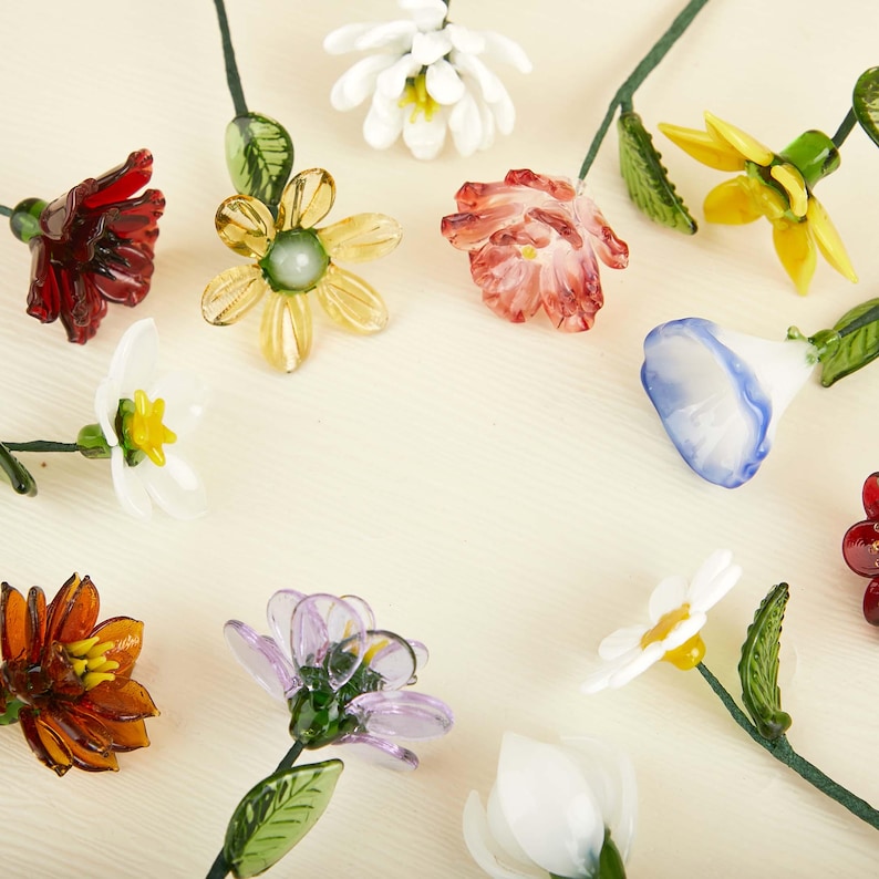 May include: A collection of handcrafted glass flowers in various colors including red, yellow, white, blue, and purple. Each flower features delicate petals, a green stem, and textured green leaves, arranged in a circular pattern on a light surface.