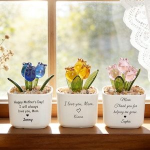 May include: Three white ceramic pots with glass tulip flowers in blue, yellow, and pink. Each pot has a message for Mother's Day, with names Jenny, Kiana, and Sophia. The pots sit on a wooden surface near a window.