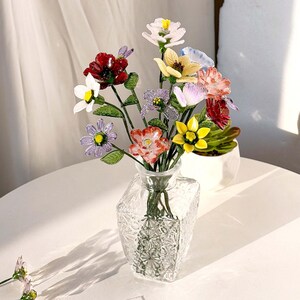 May include: A clear glass vase with a geometric textured pattern holds a bouquet of multi-colored glass flowers. The arrangement features individual stems with petals in shades of red, white, yellow, purple, and pink, accented by green leaves.