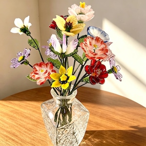 May include: A bouquet of multicolored glass flowers with green glass leaves and stems arranged in a clear, square-shaped cut glass vase. The flowers feature various shapes and colors including red, yellow, white, purple, and blue petals.