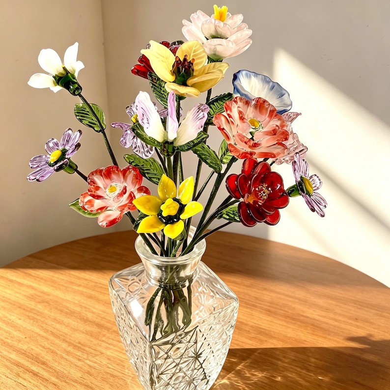 May include: A bouquet of multicolored glass flowers with green glass leaves and stems arranged in a clear, square-shaped cut glass vase. The flowers feature various shapes and colors including red, yellow, white, purple, and blue petals.