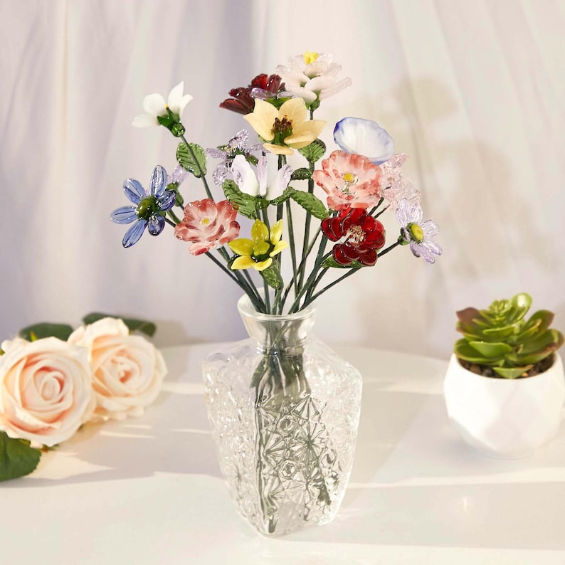 May include: A bouquet of multicolored glass flowers with green stems arranged in a clear, faceted glass vase. The flowers feature various shapes and colors including red, blue, yellow, white, and pink petals, all crafted with a glossy finish.