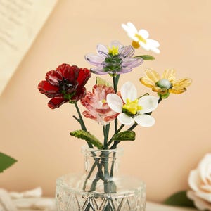 May include: A collection of small, colorful glass flowers with green stems arranged in a clear, textured glass vase. The bouquet features individual blossoms in shades of deep red, light purple, soft pink, bright white, and translucent yellow.
