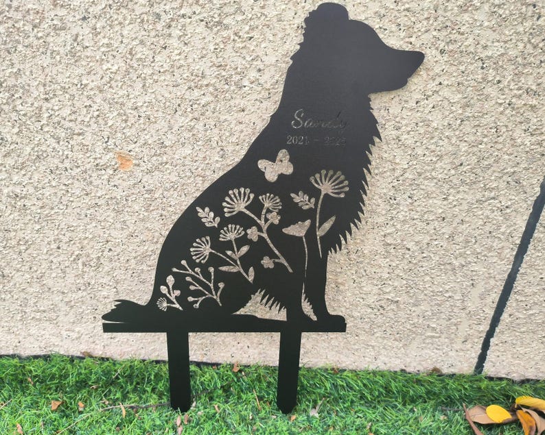 May include: Black metal silhouette of a dog, designed as a garden stake. The dog is decorated with floral and butterfly cutouts. The name "Sarah" and dates "2021-2022" are etched into the design. The stake is placed in green grass.