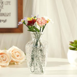 May include: A clear glass vase with a textured diamond pattern on the body, containing a small bouquet of artificial flowers in shades of purple, yellow, dark red, light pink, and white, each featuring green stems and leaves.