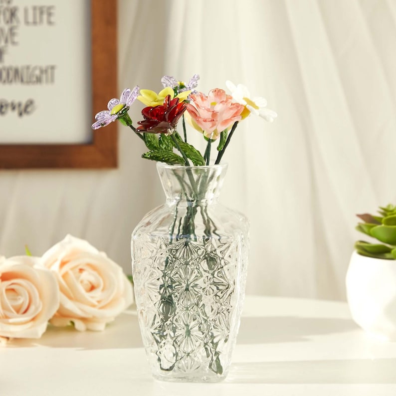 May include: A clear glass vase with a textured diamond pattern on the body, containing a small bouquet of artificial flowers in shades of purple, yellow, dark red, light pink, and white, each featuring green stems and leaves.