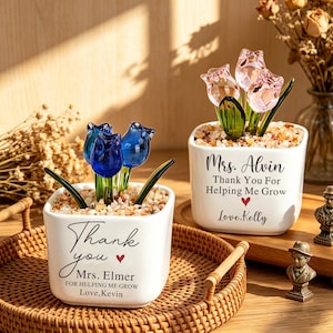 May include: Two white square ceramic planters containing glass tulips and decorative stones. One planter features blue glass tulips with text reading Thank you Mrs. Elmer for helping me grow Love, Kevin. The other has pink glass tulips with text reading Mrs. Alvin Thank You For Helping Me Grow Love, Kelly.