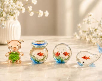 以下が含まれることがあります： 透明なガラス製の置物4点セット：四つ葉のクローバーを持つ豚、赤い魚が入った小さな丸い金魚鉢、赤い魚と緑の植物が入った球形のペーパーウェイト、そして赤い魚が入ったクジラの形をしたガラス細工のコレクション。