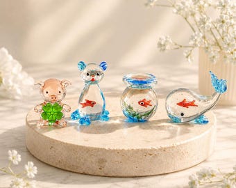 Może przedstawiać: Kolekcja czterech miniaturowych szklanych figurek, w tym świnka trzymająca zieloną czterolistną koniczynę, kot z małą pomarańczową rybką w środku, okrągła kula z pomarańczową rybką oraz wieloryb z niebieskim ogonem i pomarańczową rybką.