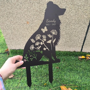 Puede incluir: Estaca de jardín de metal negro con forma de perro, con diseños florales y de mariposas. El nombre "Sandy" y los años "2021-2025" están inscritos. La estaca se sostiene con la mano y se coloca en césped verde.