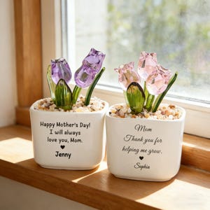以下が含まれることがあります： ガラスのチューリップの花が入った白い陶器の鉢が2つ。片方の鉢には紫色のチューリップ、もう片方にはピンク色のチューリップが入っています。鉢には「母の日おめでとう！いつもあなたを愛しています、お母さん。ジェニー」と「お母さん、私を成長させてくれてありがとう。ソフィア」と書かれています。