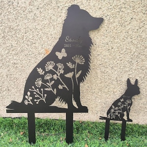 May include: Black metal dog silhouette garden stakes. The larger stake features a sitting dog with floral and butterfly cutouts, and the name "Sandy" and dates. A smaller stake depicts a dog with floral cutouts and the name "George".