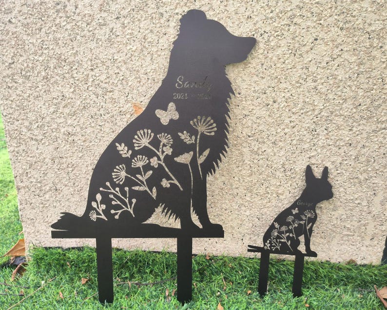 May include: Black metal dog silhouette garden stakes. The larger stake features a sitting dog with floral and butterfly cutouts, and the name "Sandy" and dates. A smaller stake depicts a dog with floral cutouts and the name "George".