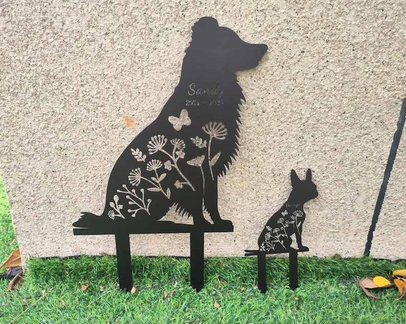May include: Black metal garden stakes featuring a dog and a rabbit silhouette. The dog stake has the name "Sandy" and dates "2021-2023" etched into it. Both stakes are decorated with floral and butterfly cutouts.
