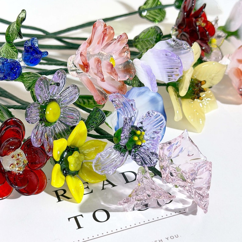May include: A collection of handcrafted glass flowers in various colors including red, yellow, blue, purple, and pink. Each flower features delicate petals and green glass stems. The flowers are arranged over a white card with the text READY TO GO.