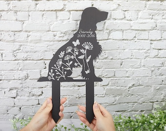 May include: Black metal garden stake in the shape of a dog, with floral and butterfly cutouts. The name "Sandy" and the years "2007-2016" are inscribed on the dog. The stake is held by two hands in front of a white brick wall.