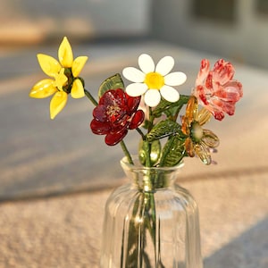 May include: A clear glass vase containing a bouquet of handcrafted glass flowers. The arrangement features a bright yellow flower, a white daisy with a yellow center, a deep red flower, a translucent amber flower, and a pinkish-red flower.