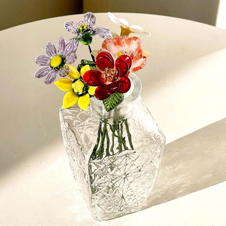 May include: A clear, square glass vase with a textured geometric pattern holds a bouquet of small, handcrafted glass flowers. The arrangement features purple, yellow, red, and white blossoms with green glass stems, creating a vibrant display.