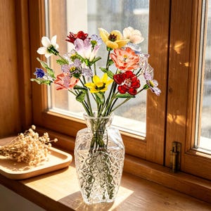 May include: A clear glass vase with a diamond-cut geometric pattern holds a bouquet of multi-colored glass flowers. The arrangement features individual blossoms in shades of red, yellow, orange, purple, blue, and white, each with green stems.