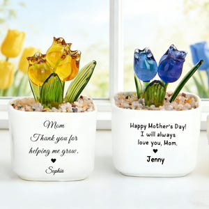 May include: Two white ceramic pots with glass flower sculptures. One pot has yellow flowers and the text "Mom, Thank you for helping me grow, Sophia." The other has blue flowers and reads "Happy Mother's Day! I will always love you, Mom, Jenny."