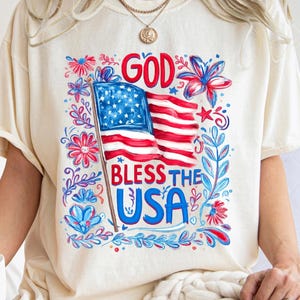May include: An ivory short-sleeve t-shirt featuring a graphic print of an American flag with the text "GOD BLESS THE USA" in red and blue. The design is surrounded by red and blue floral and leaf illustrations, creating a patriotic theme.