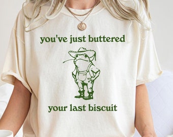 May include: A white t-shirt with a green frog wearing a hat and overalls. The text on the shirt reads "you've just buttered your last biscuit".
