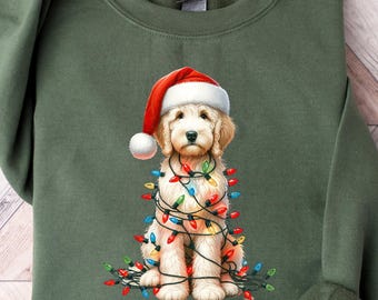 May include: An olive green sweatshirt featuring a graphic of a golden doodle wearing a Santa hat and wrapped in Christmas lights. The dog is light brown with a white face and is sitting down. The sweatshirt has long sleeves.