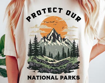 May include: Ivory-colored t-shirt with a graphic design that reads "PROTECT OUR NATIONAL PARKS." The design features a mountain range, trees, a setting sun, and flying birds. The shirt is a relaxed fit.