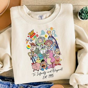 May include: Cream-colored sweatshirt featuring a colorful graphic of Toy Story characters. The design includes the words "Toy Story Land" and "To Infinity and Beyond! Est. 1995". The sweatshirt is laid on a wooden surface with a hat and jeans.