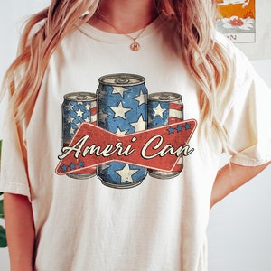 May include: A white t-shirt with a vintage-style graphic of three red, white, and blue American flag-themed cans with the text "Ameri Can" in red.