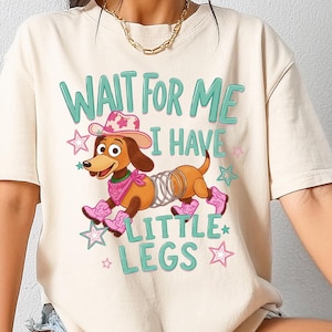 Puede incluir: Una camiseta de manga corta color crema con un gr&aacute;fico de un perro salchicha marr&oacute;n con cuerpo de resorte met&aacute;lico, que lleva un sombrero de vaquero rosa, un pa&ntilde;uelo rosa y botas de vaquero rosas, con el texto "WAIT FOR ME I HAVE LITTLE LEGS" en verde azulado.