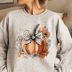May include: A light gray sweatshirt featuring a watercolor-style pumpkin design. The pumpkin is orange with a black and white polka dot bow and surrounded by floral accents in shades of orange and white. A fall-themed apparel item.