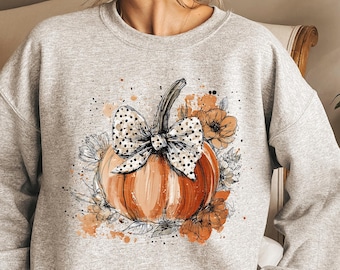 May include: A light gray sweatshirt featuring a watercolor-style pumpkin design. The pumpkin is orange with a black and white polka dot bow and surrounded by floral accents in shades of orange and white. A fall-themed apparel item.