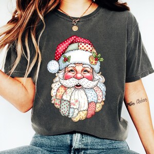 May include: Dark gray t-shirt featuring a colorful Santa Claus graphic. The Santa Claus design includes a red and white polka-dot hat, a white beard, and a patchwork design. The shirt is a comfortable, casual style.