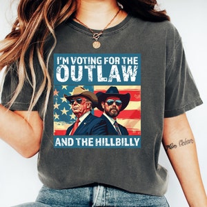 May include: A dark gray t-shirt with a distressed look featuring a graphic of two men wearing cowboy hats in front of an American flag. The text on the shirt reads "I'm voting for the Outlaw and the Hillbilly".