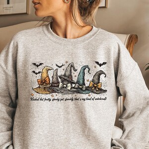 May include: A light gray sweatshirt featuring a graphic of five witch hats in various colors, each with a unique bow. Bats and stars are above the hats. The text reads "Wicked but pretty, spooky yet sparkly that's my kind of witchcraft."
