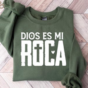 May include: A green sweatshirt with the text "DIOS ES MI ROCA" in white block letters. The word "ROCA" features a cross and a heart symbol. The sweatshirt has long sleeves and a crew neck.