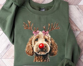 May include: A green sweatshirt featuring a dog with reindeer antlers and a red nose. The dog has brown and white fur and pink bows on the antlers. The sweatshirt has long sleeves and a crew neck.