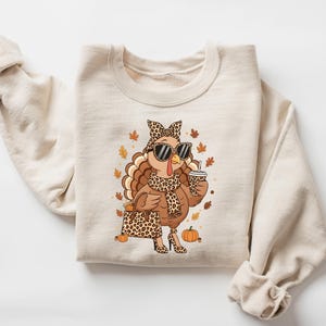 May include: Beige sweatshirt featuring a cartoon turkey wearing sunglasses, a leopard print scarf, and high heels. The turkey is holding a coffee cup and a purse, surrounded by fall leaves and pumpkins. A fun, festive design.