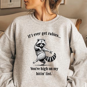 May include: A light gray sweatshirt with the text "If I ever get rabies... You're high on my bitin' list." A black and white illustration of a raccoon is featured below the text. The sweatshirt has long sleeves.