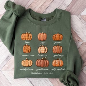 May include: An olive green crewneck sweatshirt featuring a graphic of nine pumpkins arranged in a grid. Each pumpkin is labeled with a word: love, joy, peace, forbearance, kindness, goodness, faithfulness, gentleness, self-control, and Galatians 5:22-23.