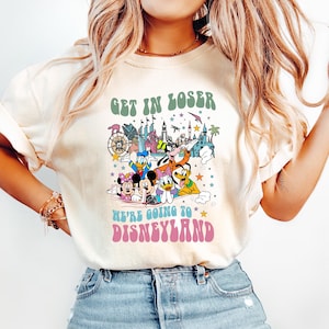 May include: Cream-colored t-shirt with the text "GET IN LOSER" above a colorful graphic of Disney characters and a castle. Below the graphic, the text reads "WE'RE GOING TO DISNEYLAND". The shirt is paired with light blue jeans.