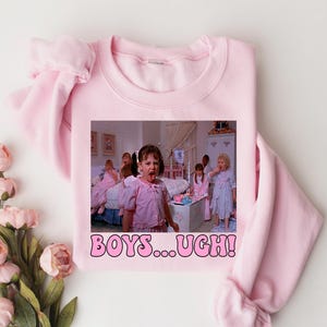 May include: A light pink sweatshirt featuring a movie scene with several girls in pajamas. The image on the sweatshirt has the text "BOYS...UGH!" in a bubble font. The sweatshirt is displayed on a white surface with pink flowers.