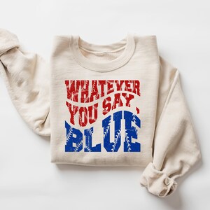 May include: Cream-colored sweatshirt with the text "WHATEVER YOU SAY, BLUE" in red and blue distressed lettering. The word "BLUE" has a baseball stitch design. The sweatshirt is folded and has rolled-up sleeves.