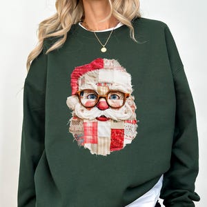 May include: Dark green sweatshirt featuring a Santa Claus face design. The Santa face is made of patchwork fabric with red, white, and beige squares. Santa wears glasses and a red hat. The sweatshirt has long sleeves and a white undershirt.